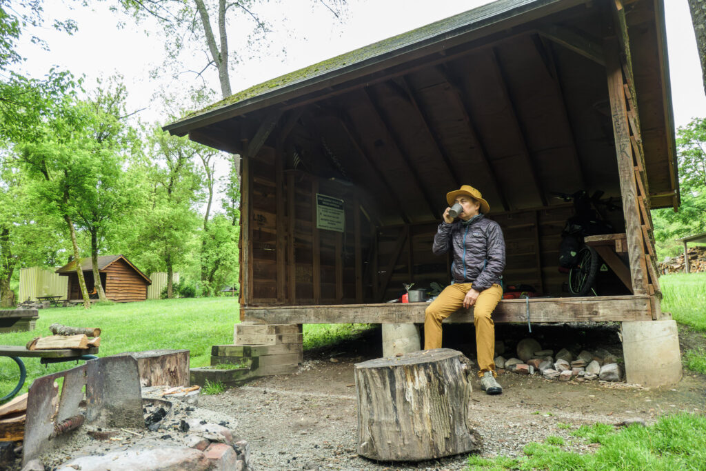A man sits at the edge of a wooden camping lean-to shelter drinking coffee. He is wearing tan pants, a purple down jacket, and a tan hat. He is looking into the distance.